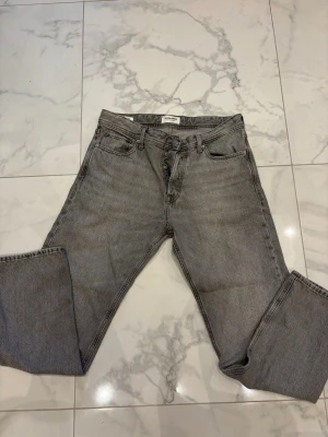 Gråa jeans från jack & Jones - Säljer ett par grå jeans från Jack & Jones med rak passform och klassisk femficksdesign. Jeansen har knappgylf och är tillverkade i ett slitstarkt denimtyg. Perfekta för en avslappnad och trendig look. 