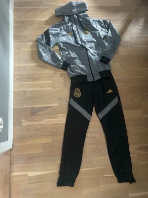 Real Madrid tränings kit  - Säljer ett snyggt träningsset från Adidas med jacka och byxor i grått och svart. Setet har guldfärgade detaljer och Real Madrid-logga på både jacka och byxor. Materialet är Climacool som andas och passar perfekt till träning eller sport.