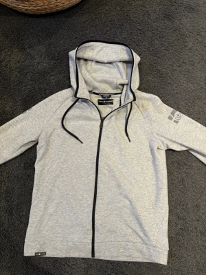 Ljusgrå sail racing hoodie med dragkedja - Snygg ljusgrå sail racing hoodie med hel dragkedja och svarta detaljer. Huva med dragsko och tryck på ena ärmen samt på huvan. Tillverkad i mjukt material som känns skönt mot huden. Perfekt för en avslappnad och sportig stil. Knappt använd och är så gott som ny.