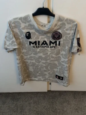 Beige camo BAPE x Miami T-shirt - Säljer en beige camo T-shirt från BAPE i samarbete med Inter Miami. T-shirten har korta ärmar, tryck fram och bak med 'MIAMI', 'A BATHING APE' och 'BAPE 93'. Skön passform och coolt mönster som sticker ut.