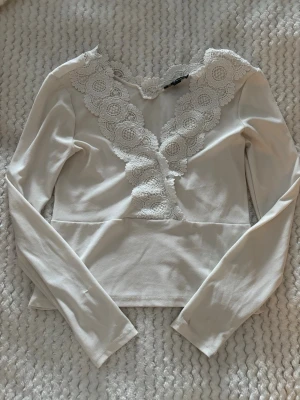 Vit blus med spetsdetaljer - Supersöt vit blus med långa ärmar och breda virkade spetsdetaljer längs v-ringningen. Blusen har en feminin och romantisk look med figurnära passform och mjukt material. Perfekt för dig som gillar detaljer och vill sticka ut lite extra.