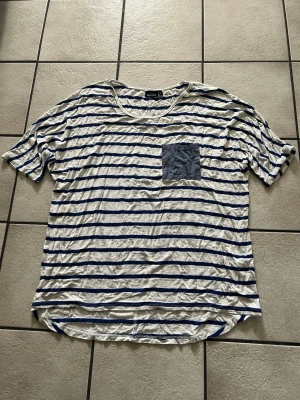 Randig t-shirt med bröstficka storlek L - Säljer en vit t-shirt med blå horisontella ränder från KappAhl. T-shirten har en rund halsringning, korta ärmar och en blå bröstficka som kontrast. Materialet är mjukt och tunt, perfekt för sommardagar. Passar dig som gillar avslappnad stil.