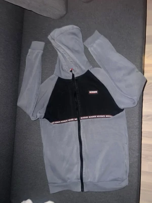 Grå hoodie med svart front och dragkedja - Säljer en grå hoodie med svart panel framtill och hel dragkedja. På bröstet finns en liten rödvit logga och ett band med texten 'McKenzie ' under den svarta delen. Hoodien har huva och är tillverkad i mjukt material, perfekt för chill dagar.