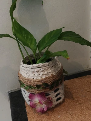 Dekorativ växt i virkad kruka - Unik grön växt i en handvirkad kruka med naturfärgad jute och vit garn. Krukan är dekorerad med ett konstgjort rosa och vitt blomster och gröna blad runt om. Perfekt som inredningsdetalj för att ge rummet en mysig känsla.