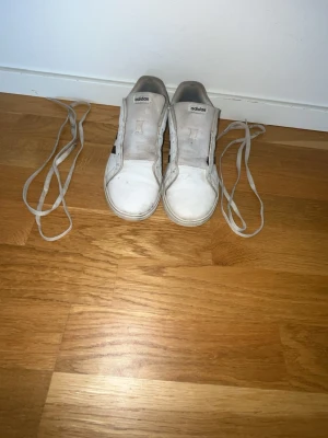 Vita Adidas sneakers med lågt skaft - Säljer ett par klassiska vita Adidas sneakers med lågt skaft. Skorna har en enkel och stilren design med vita snören och svart innersula. Tillverkade i textil och syntetmaterial, perfekta för en clean och avslappnad look. Har lite defekter som du ser på bild 2 och 5 så pris kan diskuteras.