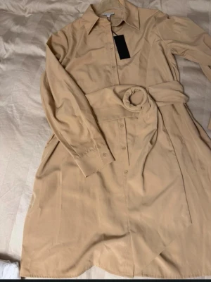 Beige skjortklänning från Gianni Bini, storlek L  - Stilren beige skjortklänning från Gianni Bini med klassisk krage, knäppning framtill och långa ärmar. Klänningen har en loose passform och är tillverkad i ett mjukt material som ger en avslappnad look. Den har ett avtagbart band i midjan för att framhäva figuren. Den kommer från rök och djurfritt hem. 