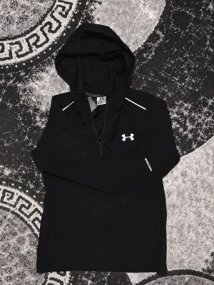 Svart windbreaker  från Under Armour - Svart hoodie från Under Armour med vit logga på bröstet och stort tryck på ryggen. Långärmad modell med huva och dragkedja framtill. Tunt och sportigt material, perfekt för träning eller chill. Snygga vita detaljer vid axlarna ger en extra touch.