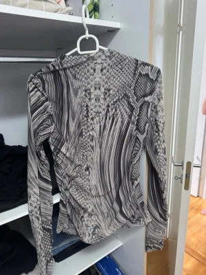 Grå långärmad topp  - Säljer en grå långärmad topp från Amisu med ett unikt ormmönster och abstrakta linjer. Toppen har en tight passform och är tillverkad i ett stretchigt syntetmaterial som känns bekvämt mot huden. Perfekt för dig som vill sticka ut med en cool look.
