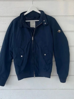 Mörkblå bomberjacka från J.Lindeberg - Snygg mörkblå jacka från J.Lindeberg i storlek xs med dragkedja framtill, 4 fickor totalt med tryckknappar och ribbade muddar. Jackan har en klassisk patch med logga på ärmen och en stilren, minimalistisk design. Perfekt för dig som gillar streetwear och enkel stil. Tveka inte att höra av dig för frågor.