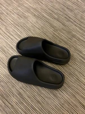 Svarta slides från Adidas - Säljer ett par svarta slides från Adidas med stilren och chunky design. Tofflorna är gjorda i ett mjukt och flexibelt syntetmaterial med öppen tå och bred rem över foten. Perfekta för chill dagar hemma eller till stranden.