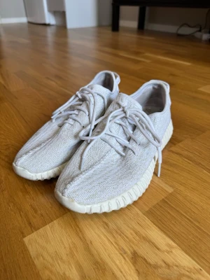 Yeezys - Säljer ett par vita sneakers med stickad ovandel och tjock, räfflad sula. Skorna har rund tå, vita skosnören och en enkel, clean design. Perfekta för dig som gillar minimalistisk stil och bekväma skor till vardags.