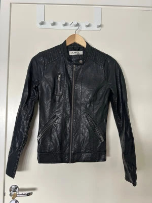 Svart skinnjacka från ONLY - Snygg svart skinnjacka från ONLY med dragkedja framtill, tre fickor med dragkedja och coola detaljer på axlarna. Jackan har en smal passform och rundad krage med tryckknapp. Perfekt för en trendig look.