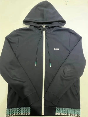 Svart hoodie med dragkedja från BOSS - Snygg svart hoodie från BOSS med vit dragkedja och huva. Diskret BOSS-logga på bröstet och upprepade BOSS-tryck längs nederkanten. Justerbara snören i huvan och ribbade muddar. Perfekt för en avslappnad och trendig stil. LIMITED EDITION HUGO BOSS SÄLJER DEN INTE MER PRIS KAN DISKUTERAS