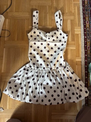 Vit prickig klänning från Zara - Supersöt vit kortklänning från Zara med stora svarta prickar. Klänningen har breda axelband, markerad midja och en utställd kjol som ger en lekfull look. Perfekt för dig som gillar retrovibbar och vill sticka ut lite extra.