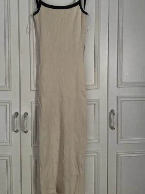 Beige långklänning med svarta band - Långklänning i beige med smala svarta axelband och kant. Klänningen har en rak, figurnära passform och är ärmlös. Materialet ser ut att vara ribbad bomull eller liknande, vilket ger en mjuk och bekväm känsla. Perfekt för varma dagar.