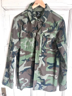 Nike SB camouflage skjorta XS - Camouflagefärgad skjorta från Nike SB i storlek XS. Skjortan har klassisk krage, två bröstfickor med knappar och är tillverkad i ett slitstarkt bomullsmaterial. Perfekt för dig som gillar streetwear och vill sticka ut med en militärinspirerad look.