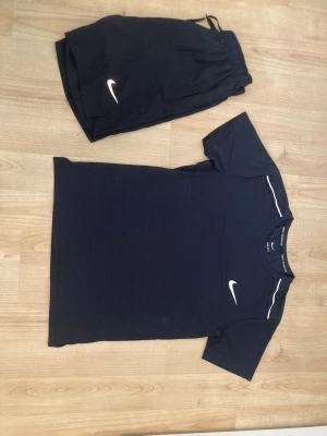 Svart Nike träningsset t-shirt & shorts - Säljer ett svart träningsset från Nike med en kortärmad t-shirt och matchande shorts. Båda plaggen har den klassiska vita Nike-loggan. T-shirten har diskreta ränder och vita detaljer vid axlarna. Perfekt för gymmet eller löpning. Storlek L