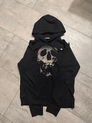 Hysteric glamour skullberry svart - Svart hoodie från Hysterics med stort dödskalletryck på framsidan och ett rött hjärta med texten 'Hysterics' på ryggen. Klassisk huva med dragsko och ribbade muddar. Perfekt för dig som gillar edgy och unik streetwear.