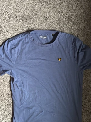 Blå t-shirt från Lyle & Scott - Snygg blå t-shirt från Lyle & Scott med klassisk rund hals och det ikoniska gula fågel-logotypen broderad på bröstet. Tillverkad i mjuk bomull och har en normal passform som funkar till det mesta.