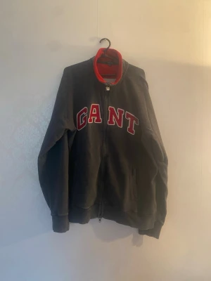 Svart zip hoodie från GANT - Snygg svart hoodie från GANT med dragkedja framtill och stora röda bokstäver på bröstet. Kragen är röd på insidan för en cool kontrast. Hoodien har ribbade muddar och två fickor fram. Perfekt för en avslappnad streetwear-look.
