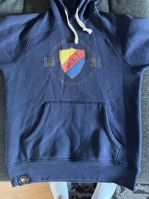Blå Djurgården hoodie med tryck - Mörkblå hoodie med stor ficka framtill och vit dragsko i huvan. På bröstet finns Djurgårdens IF:s klubbmärke och texten 'Djurgårdens Fotboll 1891 Stockholms Stolthet'. Materialet är mjukt och skönt, perfekt för chill dagar. Lite mindre i storlek mer som en medium 