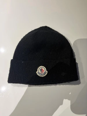 Moncler mössa - Säljer en svart ribbstickad mössa från Moncler med uppvikt kant och den klassiska Moncler-loggan framtill. Perfekt för dig som vill ha en stilren och trendig accessoar till vintern. Materialet känns mjukt och värmande.
