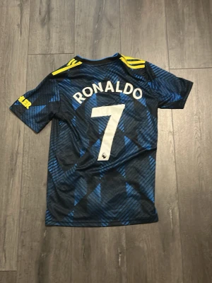 Manchester United Ronaldo matchtröja - Snygg Manchester United matchtröja från Adidas med Ronaldo och nummer 7 på ryggen. Tröjan är svart med blått mönster, gula detaljer på axlarna och klubbmärke på bröstet. Perfekt för fotbollsfans som vill sticka ut på planen eller läktaren.