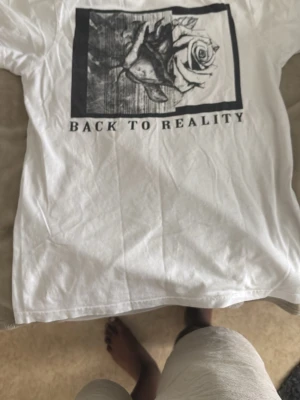 Vit t-shirt med tryck från SMOG - Vit t-shirt från SMOG i regular fit med svartvitt tryck av en ros och texten 'BACK TO REALITY' på bröstet. Klassisk rund hals och korta ärmar. Perfekt för dig som gillar stilrena plagg med coolt motiv.