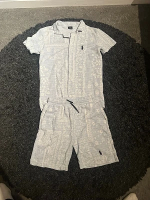 Ljusgrått set med mönster och krage - Säljer ett ljusgrått tvådelat set med kortärmad skjorta och shorts. Setet har ett diskret paisleymönster och skjortan har krage samt knappstängning. Shortsen har resår i midjan och sidofickor. Liten broderad logga på både skjorta och shorts.