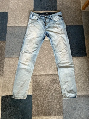 Replay jeans - Säljer ett par ljusblå replay jeans med slitningar på låret och knät. Jeansen har fem fickor, knappgylf och klassisk passform med smala ben