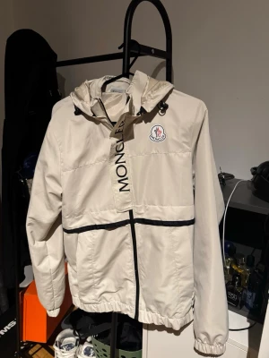 Monclizzy Vindjacka - Snygg beige vindjacka från Monclizzy med svart dragkedja och svarta detaljer. Jackan har huva med dragsko, elastiska muddar och Monc-logga på bröstet. Perfekt för blåsiga dagar och enkel att matcha med olika outfits.