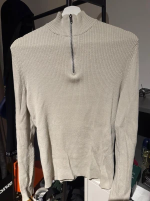 Stickad halv zip tröja - Säljer en beige ribbad stickad tröja från pier one med hög krage och dragkedja framtill. Tröjan har lång ärm och är tillverkad i ett mjukt, stretchigt material som sitter snyggt på kroppen. Perfekt för lager på lager och enkel att matcha.