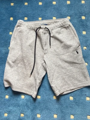 Grå sweatshorts från Polo Ralph Lauren - Snygga grå sweatshorts från Polo Ralph Lauren med svart snörning i midjan och klassisk logga broderad på vänster ben. Shortsen har två sidofickor och en bakficka, perfekta för chill dagar. Mjuk bomullskänsla och relaxed fit. De är i bra skick och passformen är S/M