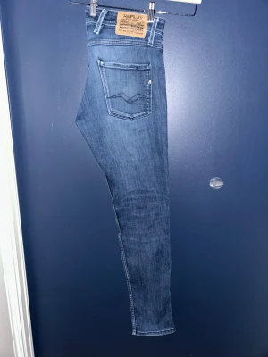 Blå slim fit jeans från Replay W30 L32 - Snygga blå slim fit jeans från Replay med klassisk femficksdesign och diskreta slitningar. Jeansen har en skön stretchig denimkvalitet och detaljerade sömmar på bakfickorna. Perfekta för en trendig och avslappnad look.