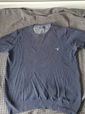 Blå stickad tröja från Gant - Snygg blå stickad tröja från Gant med rund halsringning och diskret logga broderad på bröstet. Tröjan har korta ärmar och ribbade muddar vid ärmslut och nederkant. Perfekt för en stilren och avslappnad look. Den är i rätt dåligt skick därav priset. Det går att fixa enkelt. Xs/s