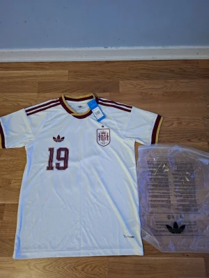 Vit Spanien fotbollströja VM 26/27 - Snygg vit fotbollströja från Adidas med Spaniens landslagsemblem och nummer 19 på framsidan. Tröjan har vinröda detaljer, klassiska Adidas-ränder på axlarna och gul/vinröd ribbad krage. Tillverkad i lätt och funktionellt material, perfekt för träning eller match.