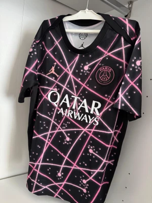 PSG x Jordan svart/rosa matchställ - Den passar för både S och M. Säljer ett PSG x Jordan matchställ med tröja och shorts. Svart bas med rosa grafiskt mönster och vita detaljer. Tröjan har PSG-logga och Jordan-logga på bröstet samt Qatar Airways-tryck. Shortsen har elastisk midja och matchande design. Perfekt för fotboll eller träning.