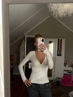 Vit omlottopp med lång ärm - Säljer en vit omlottopp med lång ärm och djup v-ringning. Toppen är tight och har en snygg omlottdesign framtill som ger en smickrande siluett. Perfekt att styla med jeans eller kjol för en clean look.