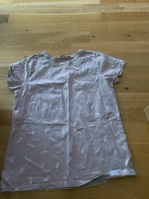 Ljusrosa t-shirt med fjädermönster - Säljer en ljusrosa t-shirt från 157 i storlek M. T-shirten har korta ärmar och är dekorerad med ett diskret vitt fjädermönster över hela plagget. Tillverkad i mjuk bomull, perfekt för en avslappnad stil.