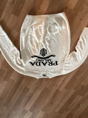Vit stickad tröja från Prada - Snygg vit stickad tröja från Prada med svart logga och texten 'PRADA MILANO' på bröstet. Tröjan har rund halsringning, långa ärmar och ribbade muddar vid ärmslut och nederkant. Materialet känns mjukt och av hög kvalitet.