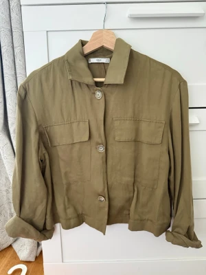 Olivgrön overshirt med fickor - Stilren olivgrön overshirt med raka linjer och två stora bröstfickor. Overshirten har krage, knäppning framtill med fyra knappar och långa ärmar som kan vikas upp. Tillverkad i ett mjukt, tunt tyg som ger en avslappnad look.