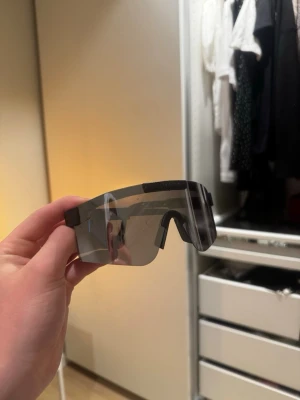 Svarta sportiga solglasögon - Säljer ett par svarta solglasögon med sportig och futuristisk design. Glasögonen har stora, rektangulära linser i mörktonat glas och breda svarta bågar. Perfekta för en cool och modern look.