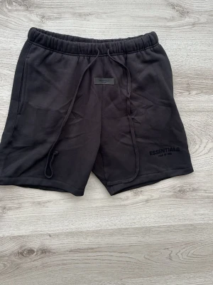  SVARTA ESSENTIALS SHORTS - Svarta shorts från Essentials Fear of God med elastisk midja och långa snören framtill. Tillverkade i mjuk bomull och har diskret logga på vänster ben. Perfekta för en avslappnad och stilren look.
