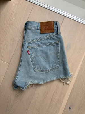Levi's ljusblå jeansshorts med fransar - Säljer ett par klassiska ljusblå jeansshorts från Levi's med råa fransade kanter och bakficka. Shortsen har hög midja och den ikoniska Levi's-lappen bak. Perfekta för sommaren och ger en avslappnad look.