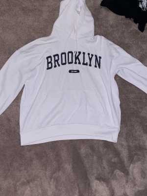 Vit hoodie BROOKLYN Iconic L - Snygg vit hoodie från Iconic Sportswear i storlek L med stor BROOKLYN-text i svart på bröstet. Klassisk känguruficka framtill, dragsko i huvan och mjukt, fleeceliknande material på insidan. Perfekt för dig som gillar streetwear och enkel stil.