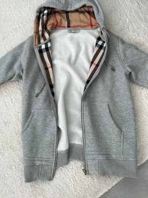 Burberry kofta - Säljer en grå zip-hoodie med klassiskt rutigt foder i huvan. Hoodien har dragkedja framtill, två fickor och ribbade muddar. Insidan av huvan har ett beige, svart och rött rutmönster som ger en snygg kontrast. Perfekt för en chill och stilren look. Oanvänd 