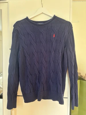 Mörkblå kabelstickad tröja Polo Ralph Lauren - Kabelstickad mörkblå tröja från Polo Ralph Lauren med klassisk röd logga broderad på bröstet. Tröjan har rund halsringning, långa ärmar och ribbade muddar. Perfekt för en stilren och avslappnad look.