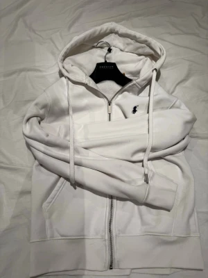 Vit hoodie från Polo Ralph Lauren - Säljer en stilren vit hoodie från Polo Ralph Lauren med dragkedja framtill och klassisk blå logga på bröstet. Hoodien har huva med dragsko, ribbade muddar och fickor på sidorna. Perfekt för en clean och avslappnad look.