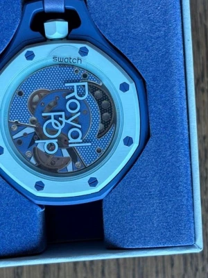 Blå Royal Pop klocka Swatch x AP - Säljer en blå Royal Pop Lan Ba klocka från Swatch och Audemars Piguet. Klockan har ett genomskinligt urverk med blå detaljer och texten 'Royal Pop' på urtavlan. Snygg oktagonal form och matchande blått armband. Perfekt för dig som gillar unika accessoarer.