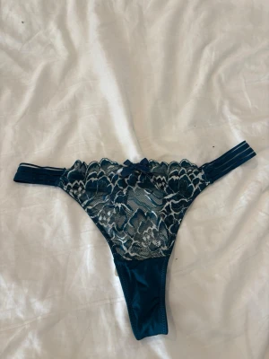 Mörkblå oanvända spetsstring med rosett från hunkemöller - Snygg mörkblå stringtrosa med breda sidoband och detaljerad spets framtill. Dekorerad med en liten rosett i midjan och genomskinliga partier i spetsen. Materialet är en mix av spets och mjuk stretchig textil för bekväm passform. Oanvända med lappen kvar 